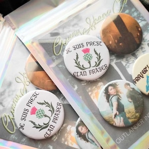 May include: Four round button pins with different designs. One pin features a white background with the text "Je suis prest Clan Fraser" and a green thistle. Another pin features a photo of a woman and man in a field. The third pin features a photo of a building. The fourth pin features a white background with the text "Fraser" in pink.