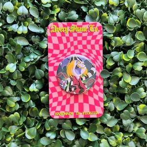May include: A round button with a colorful illustration of a character with a green and black striped shirt, a red bow tie, and a white face with a wide, toothy grin. The button is on a pink and white checkered background with the text "Avery Shame Co." and "Handmade in Florida".