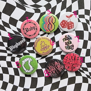 May include: Nine colorful round pins with various designs and text. The pins feature designs such as a skull, a campfire, a cherry, and a heart. The text on the pins includes "Blehgssed", "Baby", "Midnight Society Member", "Thick and Tired", "Anti Social Mommy Club", and "Baby Girl".