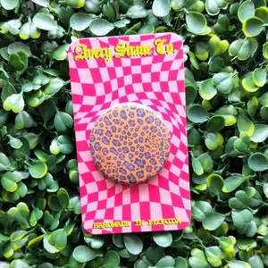 May include: A pink and white checkered background with a round, orange and purple leopard print phone grip. The phone grip is on a card that says "Avery Shane Co." and "Handmade in Florida".