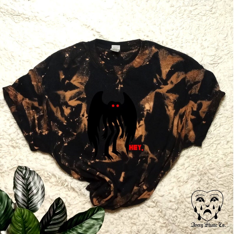 May include: Black t-shirt with a bleach tie-dye pattern. The shirt features a black silhouette of a creature with red eyes and the word "HEY." printed in red below it.