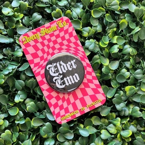 May include: A black button with the words "Elder Emo" in white gothic font, displayed on a pink and white checkered card. The card has text that reads "Avery Shane Co." and "HANDMADE IN FLORIDA". The button and card are set against a backdrop of green foliage.