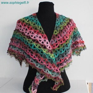 Può includere: Uno scialle all'uncinetto multicolore, esposto su un manichino nero. Lo scialle presenta un mix di filati rosa, verdi, blu e grigi, con un design traforato. Il testo "Création Sophie Gelfi Designs" è visibile. Fatto a mano.