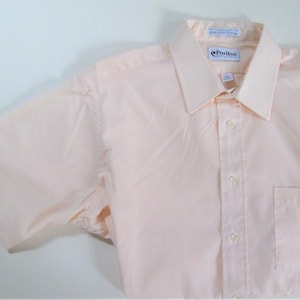 May include: A light pink short-sleeved button-down shirt with a Puritan brand label. The shirt has a pocket on the left side and a button-up front.