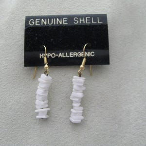 May include: A pair of white shell earrings with gold-colored hooks. The earrings are made of small, stacked shell pieces. The black tag above the earrings reads "GENUINE SHELL" and "HYPO-ALLERGENIC".