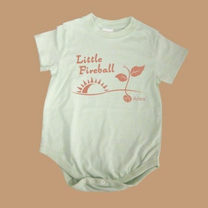 K&ouml;nnte beinhalten: Ein hellgr&uuml;ner Baby-Strampler mit einer braunen Grafik einer Sonne, die &uuml;ber einer Pflanze aufgeht, mit dem Text "Little Fireball" und "Aries" darunter.