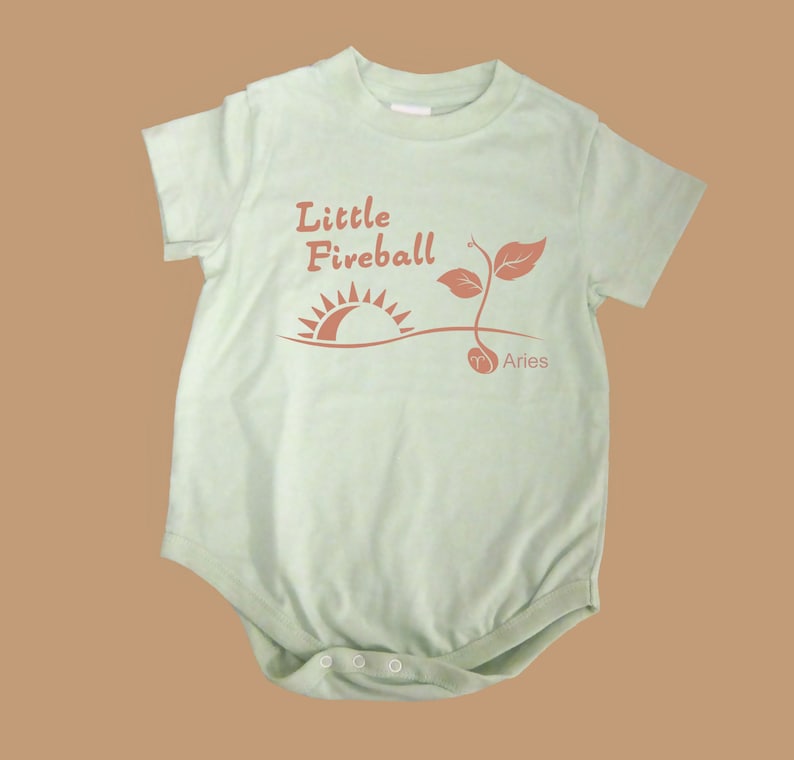 K&ouml;nnte beinhalten: Ein hellgr&uuml;ner Baby-Strampler mit einer braunen Grafik einer Sonne, die &uuml;ber einer Pflanze aufgeht, mit dem Text "Little Fireball" und "Aries" darunter.