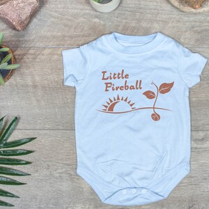 K&ouml;nnte beinhalten: Ein hellblauer Baby-Strampler mit dem Text "Little Fireball" in braunen Buchstaben. Der Strampler zeigt eine braune Sonne und einen braunen Spross mit einem Samen.