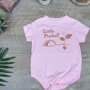 K&ouml;nnte beinhalten: Rosaer Baby-Strampler mit einer braunen Grafik eines Sonnenaufgangs und eines Sprosses mit dem Text "Little Fireball".