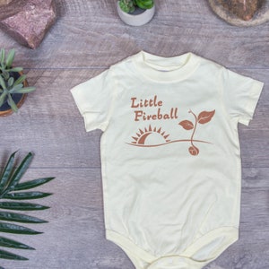 K&ouml;nnte beinhalten: Ein hellgelber Baby-Strampler mit einer braunen Grafik eines Sonnenaufgangs und eines Sprosses. Der Text "Little Fireball" ist in Braun &uuml;ber der Grafik gedruckt.