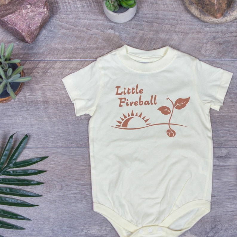 K&ouml;nnte beinhalten: Ein hellgelber Baby-Strampler mit einer braunen Grafik eines Sonnenaufgangs und eines Sprosses. Der Text "Little Fireball" ist in Braun &uuml;ber der Grafik gedruckt.