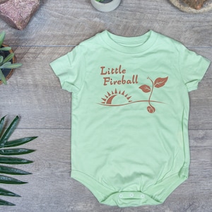 K&ouml;nnte beinhalten: Ein hellgr&uuml;ner Baby-Strampler mit dem Text "Little Fireball" in braunen Buchstaben. Der Strampler zeigt eine braune Grafik eines Sonnenaufgangs und einer kleinen Pflanze.
