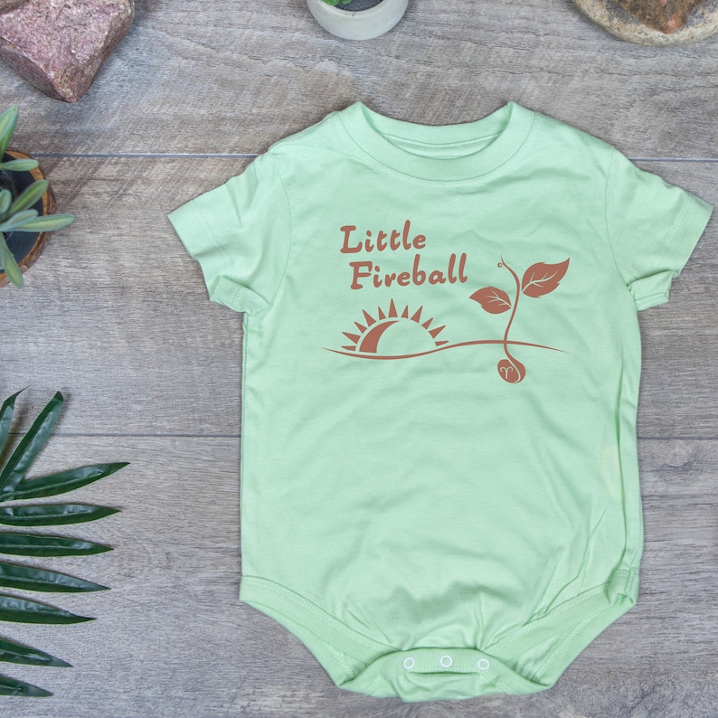 K&ouml;nnte beinhalten: Ein hellgr&uuml;ner Baby-Strampler mit dem Text "Little Fireball" in braunen Buchstaben. Der Strampler zeigt eine braune Grafik eines Sonnenaufgangs und einer kleinen Pflanze.