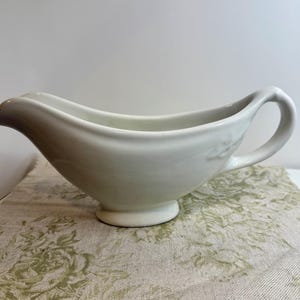 May include: White ceramic gravy boat with a handle. The boat is sitting on a floral patterned fabric.