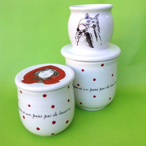 May include: Three white ceramic containers with red polka dots. The top container features a black and white illustration of a wolf. The middle container has a white lid. The bottom container has a red lid with a black and white illustration of a girl wearing a red hood. All three containers have the text "lait et un petit pot de beurre" on the side.