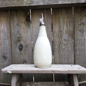 May include: A white ceramic bottle with a silver top and a spout. The bottle is sitting on a wooden stool.
