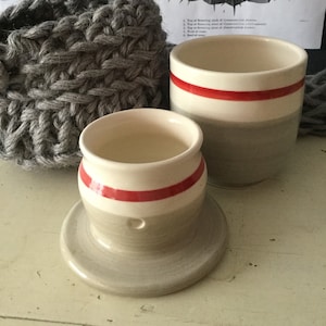 May include: Two ceramic butter dishes with red and white stripes. The smaller dish has a hole in the bottom and sits on a matching saucer. The larger dish is taller and has a wider opening.