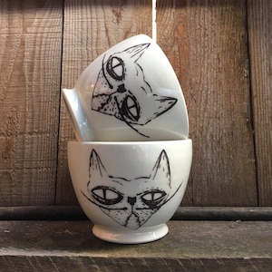 May include: Two white ceramic bowls with black hand-painted cat faces. The bowls are stacked on top of each other. The cat faces have large eyes and whiskers.