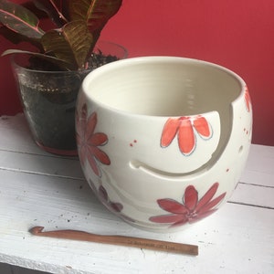Puede incluir: Cuenco de lana de cer&aacute;mica blanca con un dise&ntilde;o floral rojo y naranja. El cuenco tiene un agujero en el lateral para que la lana pueda pasar. Un gancho de ganchillo de madera est&aacute; en primer plano.