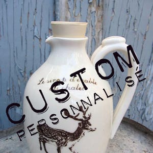 May include: Cream ceramic pitcher featuring a deer illustration and the text "Le sirop d'érable chalet". The pitcher has a handle and a cork stopper. The word "CUSTOM PERSONNALISÉ" is written on the pitcher.