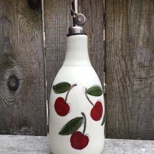 Puede incluir: Botella de aceite de oliva de cerámica blanca con un pico de vertido plateado y un diseño de cerezas. La botella tiene tres cerezas rojas con tallos y hojas verdes.