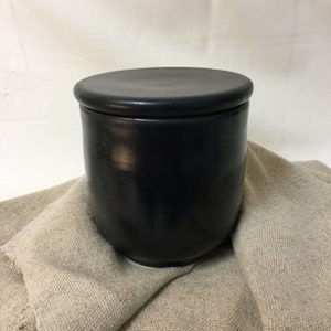 Pode incluir: Um pote de cerâmica preto com tampa a condizer. O pote tem formato arredondado e assenta sobre um pano de linho bege. O pote tem aproximadamente 10 cm de altura e 10 cm de largura.