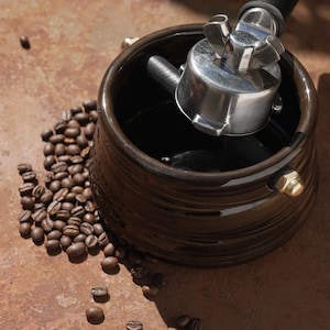Puede incluir: Una cafetera de cerámica marrón oscuro con un portafiltro de acero inoxidable y un montón de granos de café tostados. La cafetera tiene forma redonda con un exterior texturizado y detalles en latón.