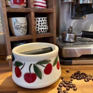 Peut inclure: Un récipient à marc de café en céramique blanche avec un motif de cerises, une barre noire et un raccord en laiton. Des grains de café sont dispersés sur la surface en bois. D'autres tasses en céramique sont en arrière-plan.