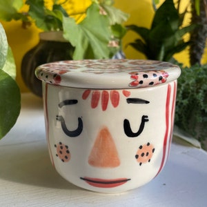 May include: A white ceramic container with a red and black patterned lid. The container has a hand-painted face with closed eyes, a pink nose, and a red mouth.