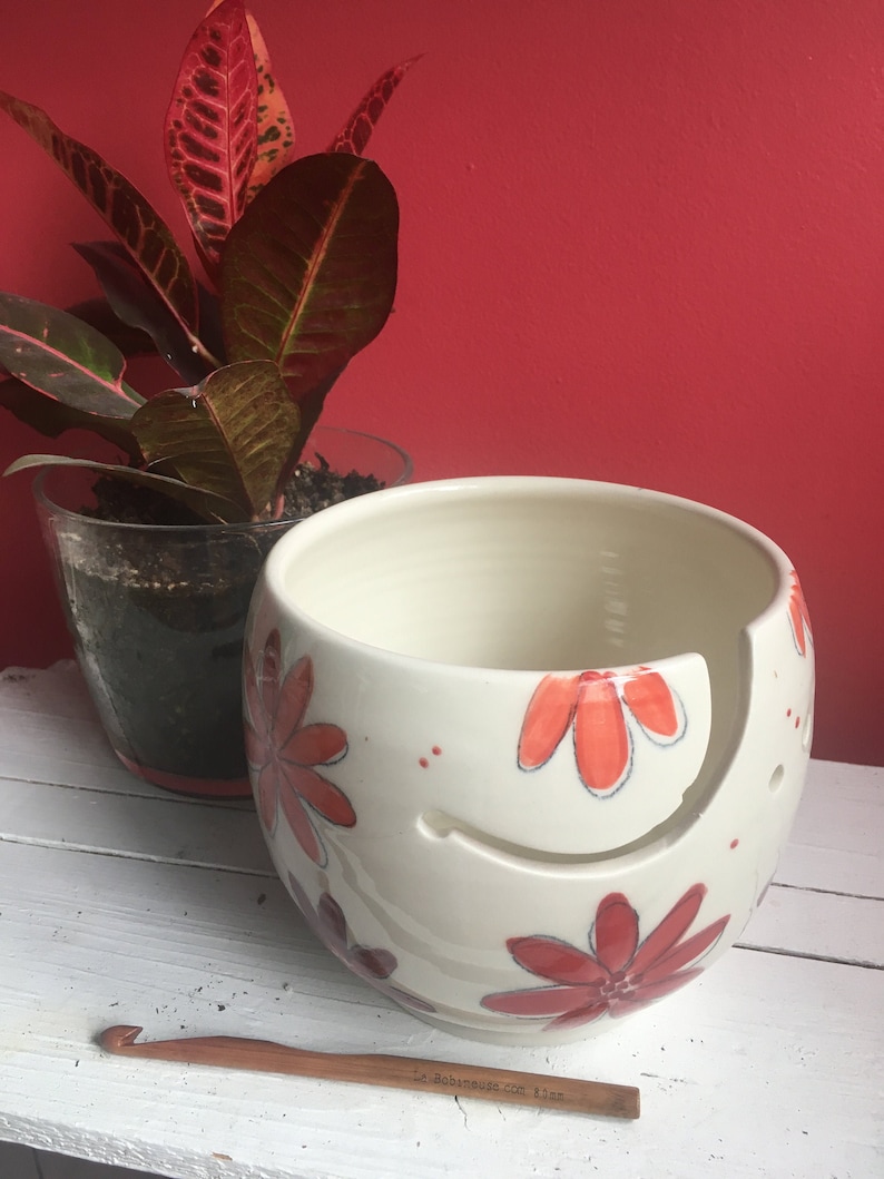 Puede incluir: Cuenco de lana de cer&aacute;mica blanca con un dise&ntilde;o floral rojo y naranja. El cuenco tiene un agujero en el lateral para que la lana pueda pasar. Un gancho de ganchillo de madera est&aacute; en primer plano.