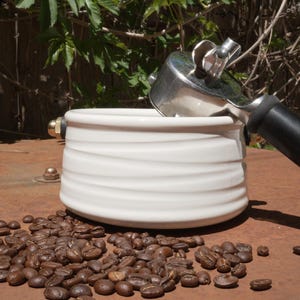 May include: A white ceramic espresso cup with a ridged design, partially filled with a silver portafilter. Roasted coffee beans are scattered around the cup on a rusty brown surface. The background features green foliage.