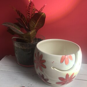 Puede incluir: Cuenco de lana de cer&aacute;mica blanca con un dise&ntilde;o floral rojo, naranja y amarillo. El cuenco tiene un hueco para que la lana pase a trav&eacute;s. Un gancho de ganchillo de madera est&aacute; en primer plano.