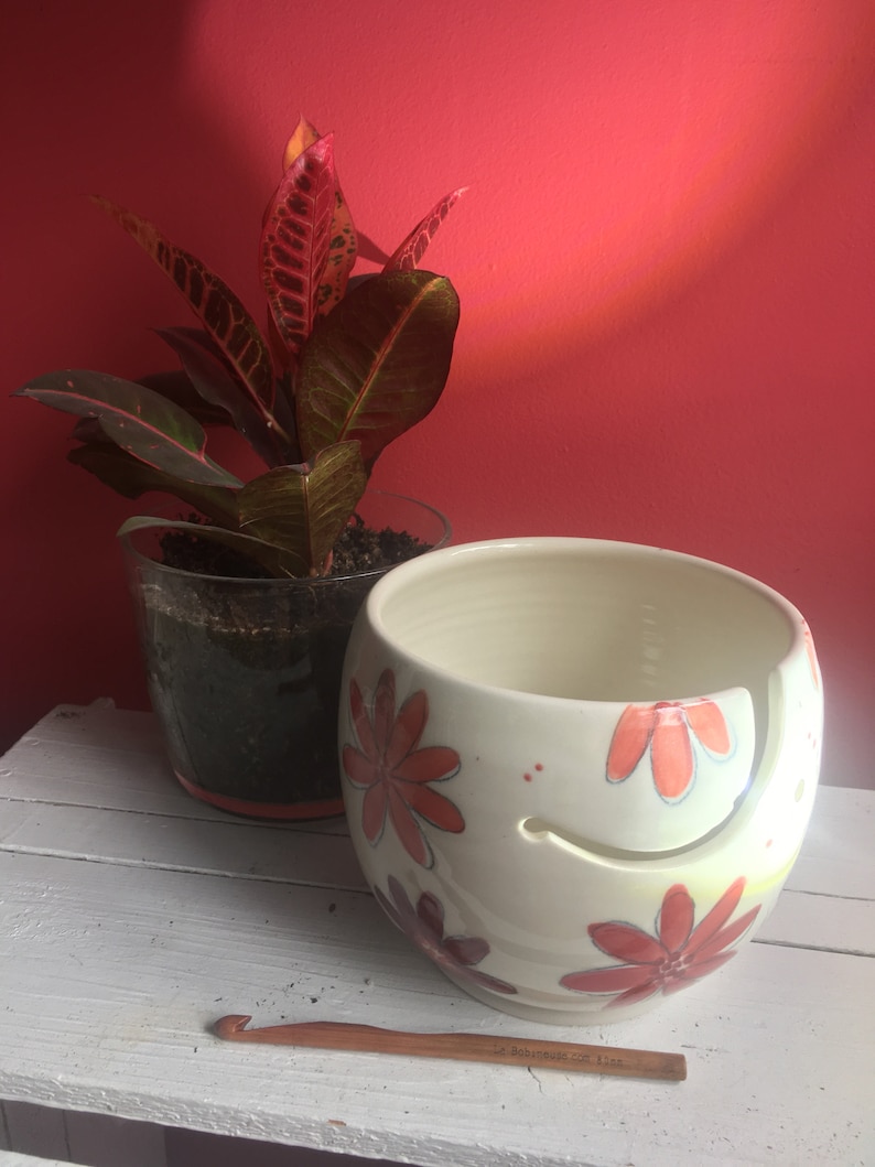 Puede incluir: Cuenco de lana de cer&aacute;mica blanca con un dise&ntilde;o floral rojo, naranja y amarillo. El cuenco tiene un hueco para que la lana pase a trav&eacute;s. Un gancho de ganchillo de madera est&aacute; en primer plano.