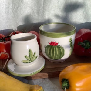 Könnte beinhalten: Zwei Keramikbehälter mit grünen und roten Kaktus-Designs. Die Behälter stehen auf einer Holzoberfläche mit gelben und roten Paprikaschoten.