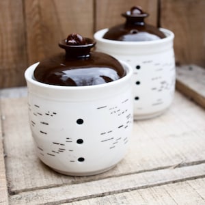 Può includere: Due barattoli di ceramica bianca con coperchi marroni e segni neri. I barattoli hanno piccoli fori sui lati.