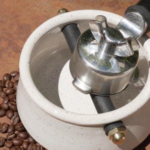May include: A white speckled ceramic coffee maker component with a shiny metal portafilter. A black tube runs through the center. Coffee beans are scattered around the base of the maker. The background is a rusty brown surface.