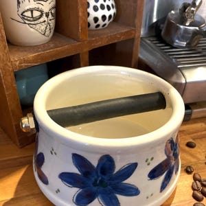 Peut inclure: Un récipient à café en céramique blanche avec une poignée noire et un motif floral bleu. Le récipient est posé sur une surface en bois.