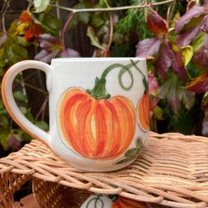 May include: A white ceramic mug with a hand-painted design of orange pumpkins and green vines. The mug has a rounded shape and a handle.