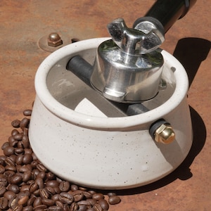 Könnte beinhalten: Ein weißes Keramik-Kaffeezubereitungsgerät mit einem Edelstahlventil und einem schwarzen Schlauch. Geröstete Kaffeebohnen sind um die Basis des Geräts verstreut. Das Gerät steht auf einer rostbraunen Oberfläche.