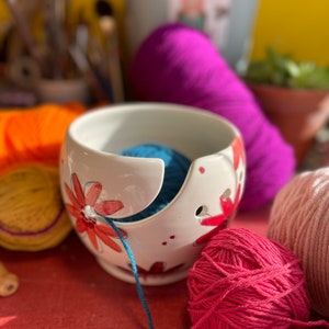 Puede incluir: Un bol de lana de cer&aacute;mica blanca con un dise&ntilde;o floral y una bola de lana azul en su interior. El bol tiene un agujero para que la lana pase a trav&eacute;s de &eacute;l. Tambi&eacute;n hay bolas de lana rosa y azul sobre una superficie roja.