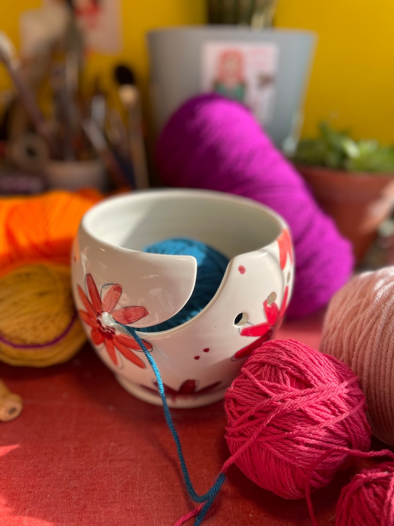 Puede incluir: Un bol de lana de cer&aacute;mica blanca con un dise&ntilde;o floral y una bola de lana azul en su interior. El bol tiene un agujero para que la lana pase a trav&eacute;s de &eacute;l. Tambi&eacute;n hay bolas de lana rosa y azul sobre una superficie roja.