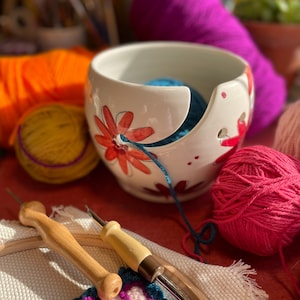 Puede incluir: Un bol de lana de cer&aacute;mica blanca con un dise&ntilde;o floral y una bola de lana azul en su interior. El bol est&aacute; sobre una superficie roja con una bola de lana rosa, una bola de lana amarilla y dos agujas de crochet de madera.