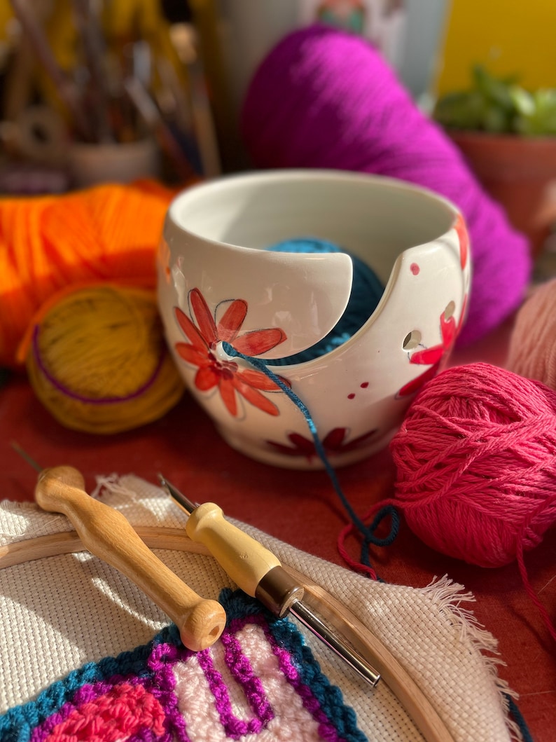 Puede incluir: Un bol de lana de cer&aacute;mica blanca con un dise&ntilde;o floral y una bola de lana azul en su interior. El bol est&aacute; sobre una superficie roja con una bola de lana rosa, una bola de lana amarilla y dos agujas de crochet de madera.