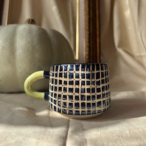 May include: A ceramic mug with a blue and white checkered pattern and a light green handle. The mug is sitting on a white fabric surface.