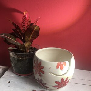 Puede incluir: Cuenco de lana de cer&aacute;mica blanca con un dise&ntilde;o floral en tonos naranja y rojo. El cuenco tiene un agujero para que la lana pase a trav&eacute;s de &eacute;l. Tambi&eacute;n se muestra un gancho de ganchillo de madera.