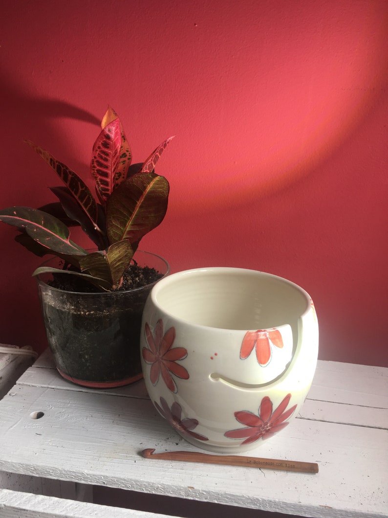 Puede incluir: Cuenco de lana de cer&aacute;mica blanca con un dise&ntilde;o floral en tonos naranja y rojo. El cuenco tiene un agujero para que la lana pase a trav&eacute;s de &eacute;l. Tambi&eacute;n se muestra un gancho de ganchillo de madera.