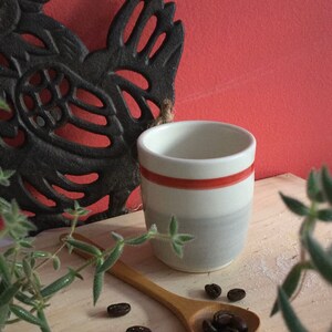 May include: A white ceramic mug with a red stripe and a grey glaze. The mug is sitting on a wooden surface with a wooden spoon and coffee beans.