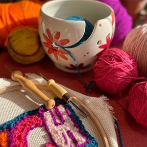 Puede incluir: Un bol de lana de cer&aacute;mica blanca con dise&ntilde;os florales y una bola de lana azul en su interior. El bol est&aacute; sobre una superficie roja con una aguja de lana de madera, un aro con un proyecto de bordado de colores y varias bolas de lana de diferentes colores.
