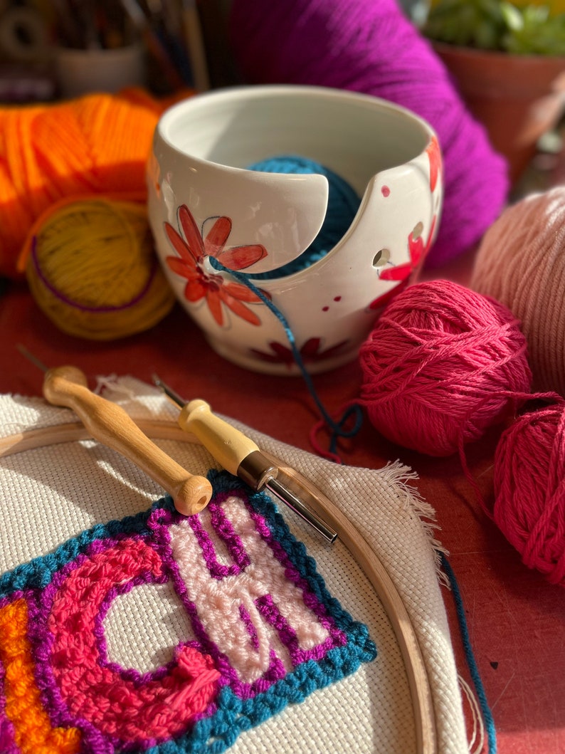 Puede incluir: Un bol de lana de cer&aacute;mica blanca con dise&ntilde;os florales y una bola de lana azul en su interior. El bol est&aacute; sobre una superficie roja con una aguja de lana de madera, un aro con un proyecto de bordado de colores y varias bolas de lana de diferentes colores.