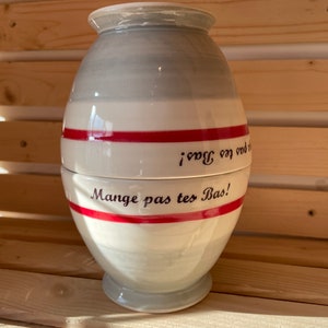 May include: A ceramic jar in light grey and white, with a red stripe and the text "Mange pas tes Bas!" in black. The jar has a rounded form and a lid, ideal for kitchen storage. The measurements are in centimetres.